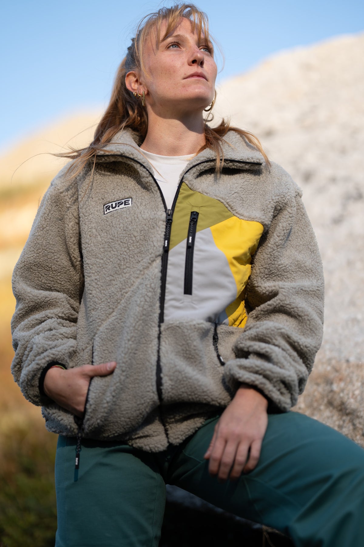 FW26-126 Yeti 2.0 Sunny + Ande Forest Green (donna seduta, guarda in alto, cielo blu)